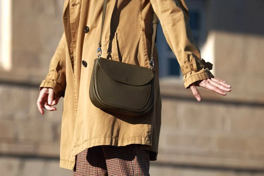 Personne portant un trench-coat beige et un pantalon à carreaux, portant un sac à bandoulière vert foncé, le bras tendu sur le côté.