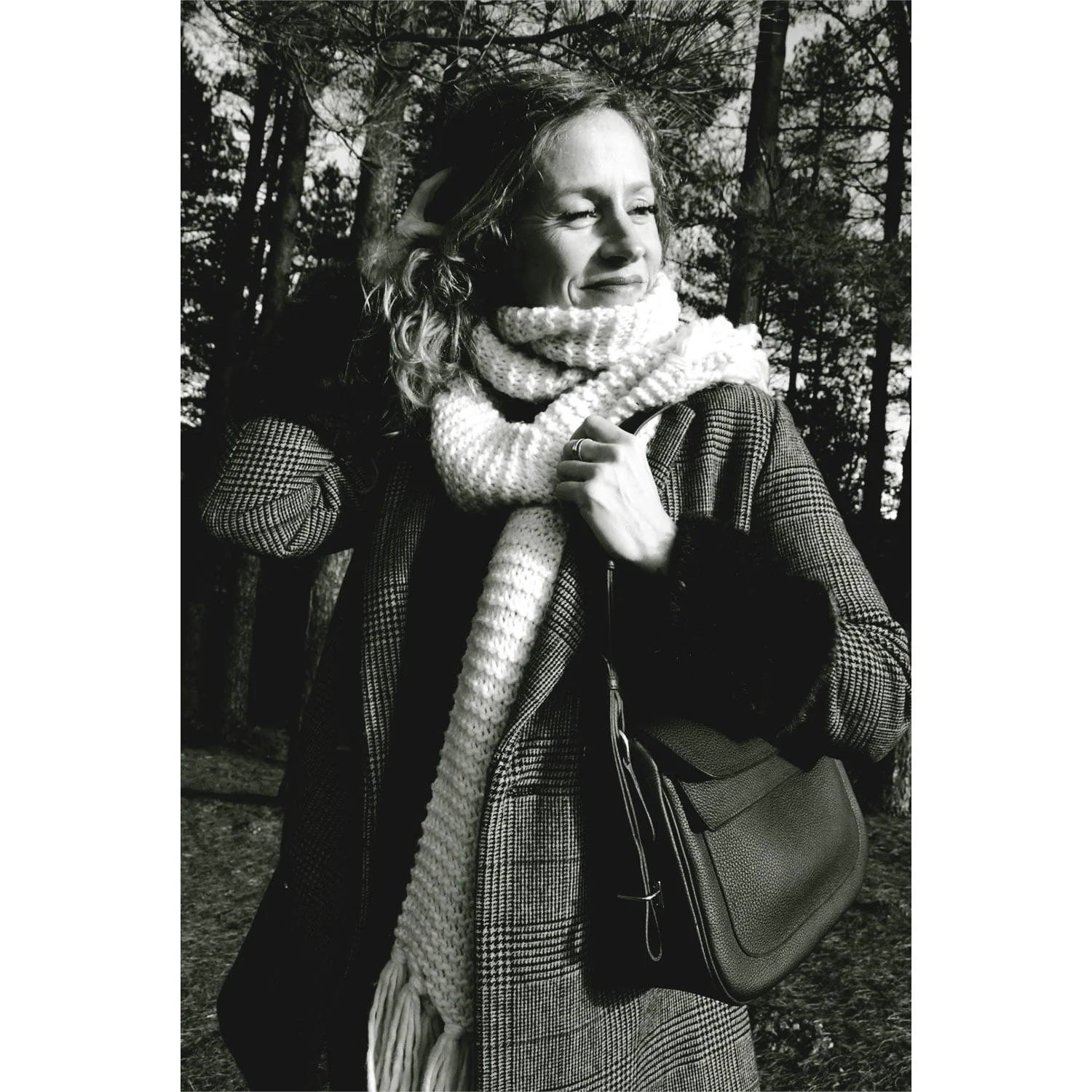 Une femme vêtue du manteau et de l'écharpe ADELE se tient debout à l'extérieur, tenant un sac, avec des arbres en arrière-plan. L'image est représentée en noir et blanc.