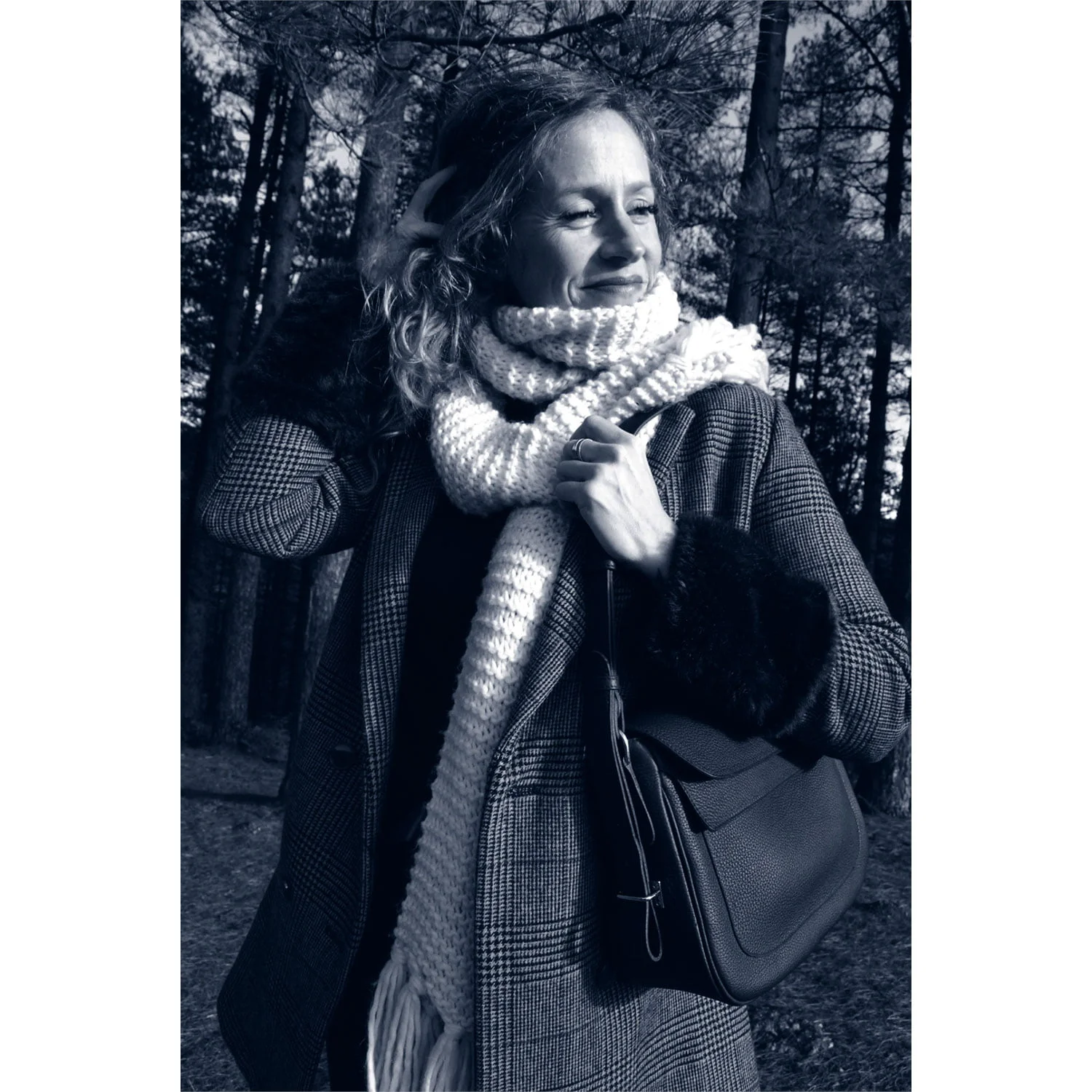 Une femme portant un manteau et une écharpe se tient dans une forêt, tenant le sac à main ADELE.