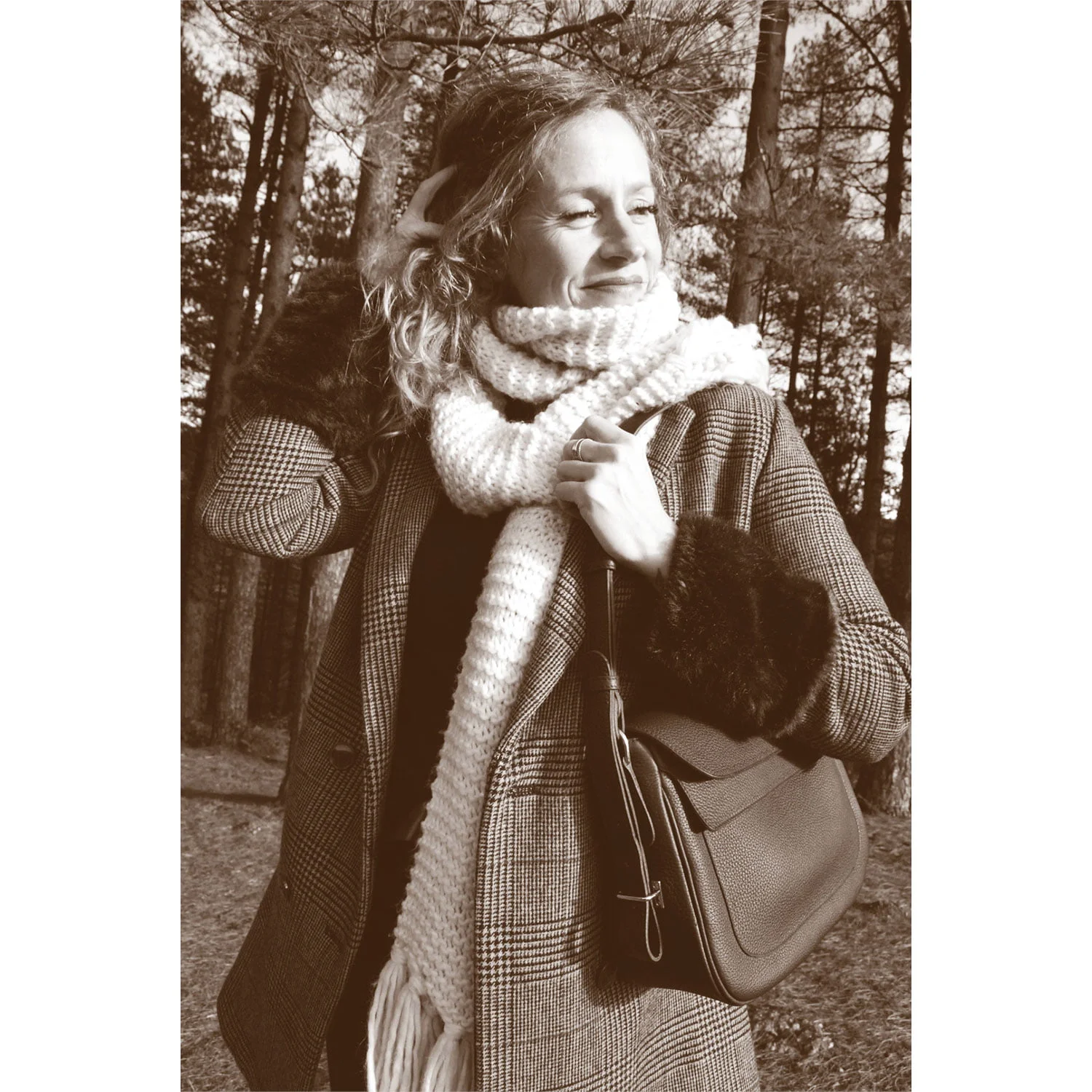 Une femme portant le manteau à carreaux ADELE et une longue écharpe se tient debout dans une forêt, souriante, une main sur la tête et l'autre tenant un sac à main. L'image a un ton sépia.