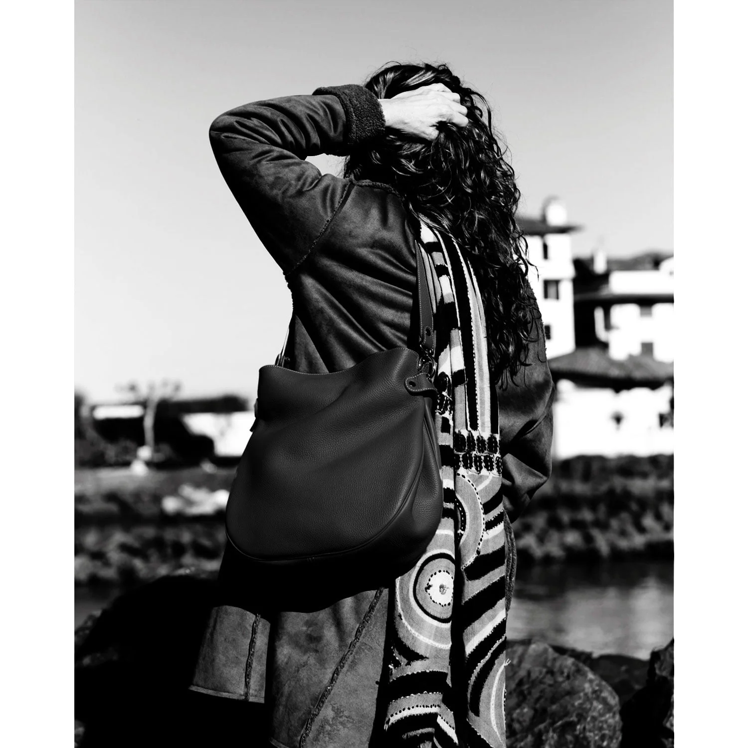 Sur une image monochrome, une personne aux cheveux longs se tient dos à la caméra, vêtue d'un manteau et d'une écharpe à motifs, tout en tenant le sac à main GABY. Ils sont à l'extérieur, encadrés par un paysage urbain et de l'eau en arrière-plan.