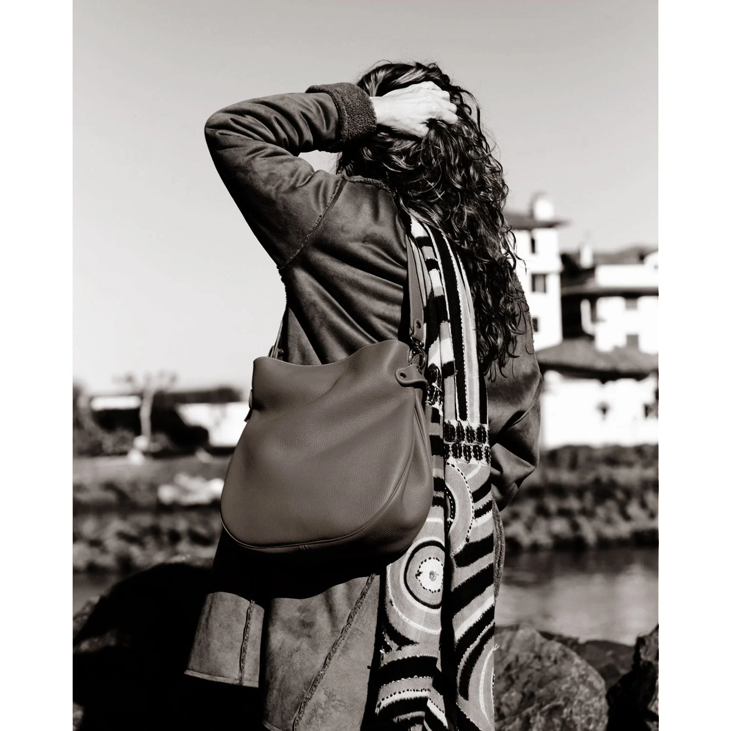 Vêtue du manteau GABY et d'une écharpe à motifs, une personne se tient près de l'eau avec le grand sac en bandoulière, regardant ailleurs.