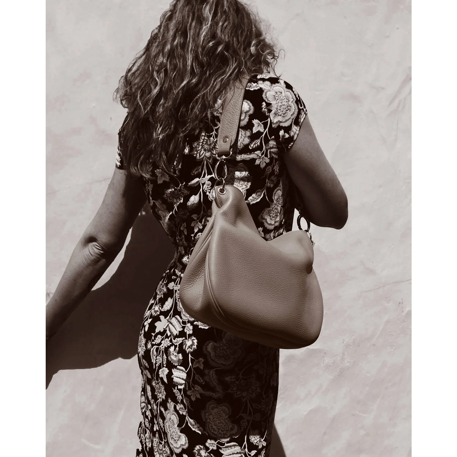 Une femme aux cheveux bouclés, vêtue d'une tenue fleurie, tient le grand sac à main JUDY contre un mur uni.
