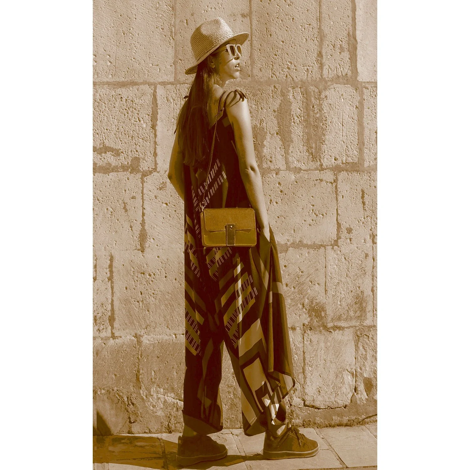 Une personne portant un chapeau, des lunettes de soleil et une robe à motifs se tient contre un mur de pierre, tenant le sac LOU FAÇON GALUCHAT.