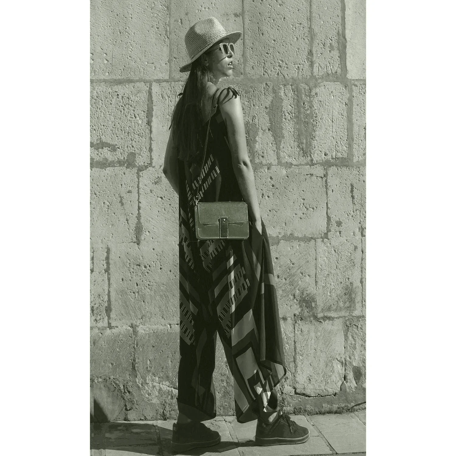 Une femme vêtue d'une robe à motifs, d'un chapeau et de lunettes de soleil se tient debout face à un mur de pierre, le dos partiellement tourné vers l'objectif. Elle porte le sac bandoulière LOU FAÇON GALUCHAT.