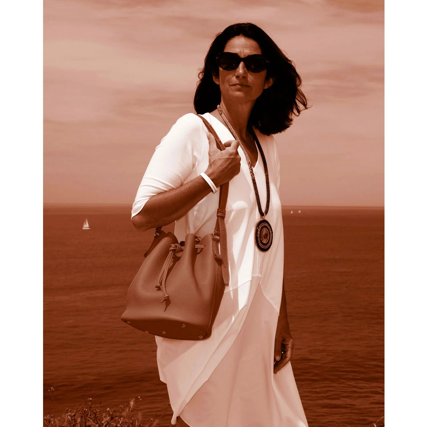 Une femme portant des lunettes de soleil et une robe blanche LOUISON se tient au bord de la mer, portant un sac à main, avec des voiliers visibles en arrière-plan.