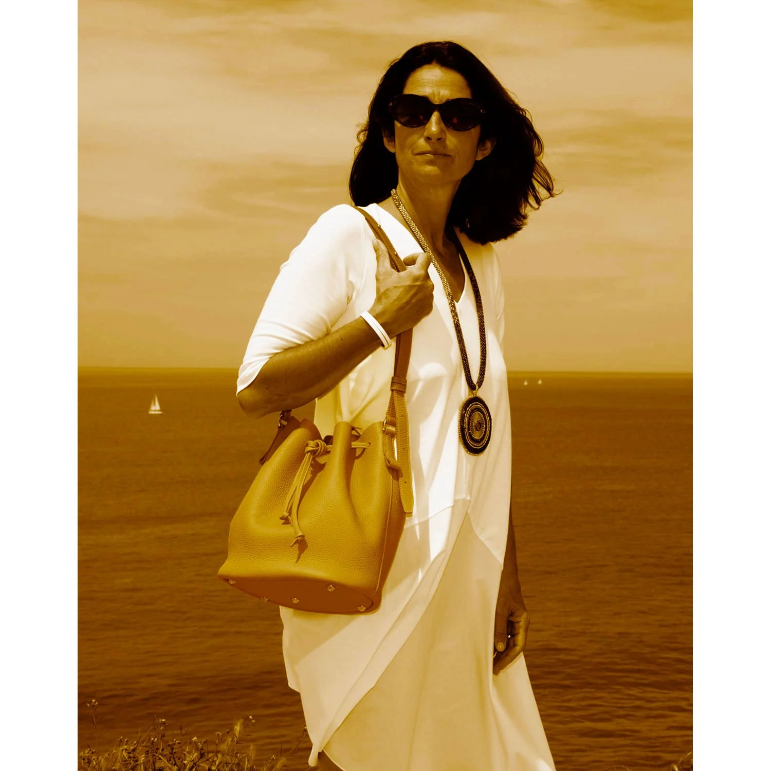 Une femme portant des lunettes de soleil, vêtue de la robe blanche LOUISON et portant un sac à main beige, se tient au bord de la mer avec des voiliers en arrière-plan sous un ciel partiellement nuageux.