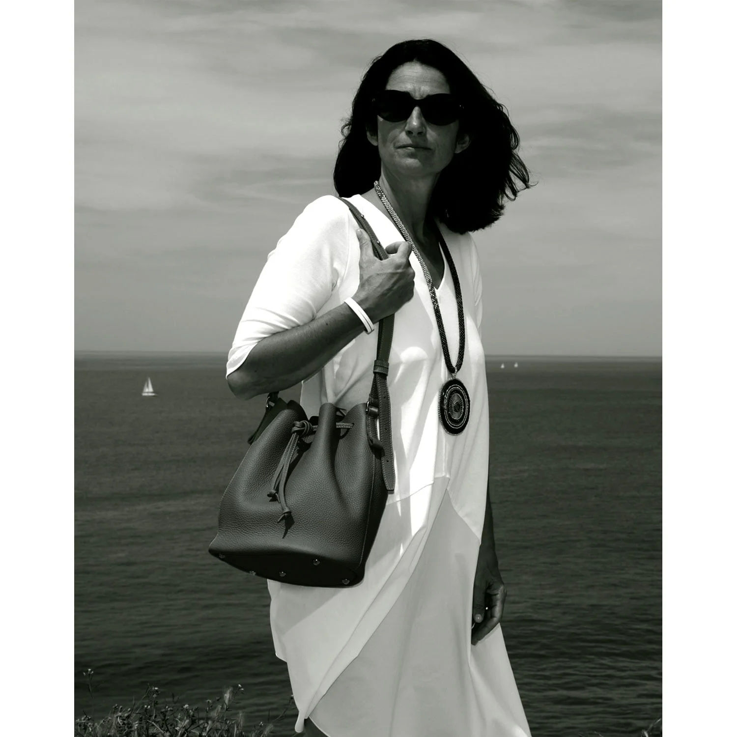 Une femme portant des lunettes de soleil et une tenue blanche se tient près de l'océan, tenant le sac à main LOUISON, avec des voiliers en arrière-plan.