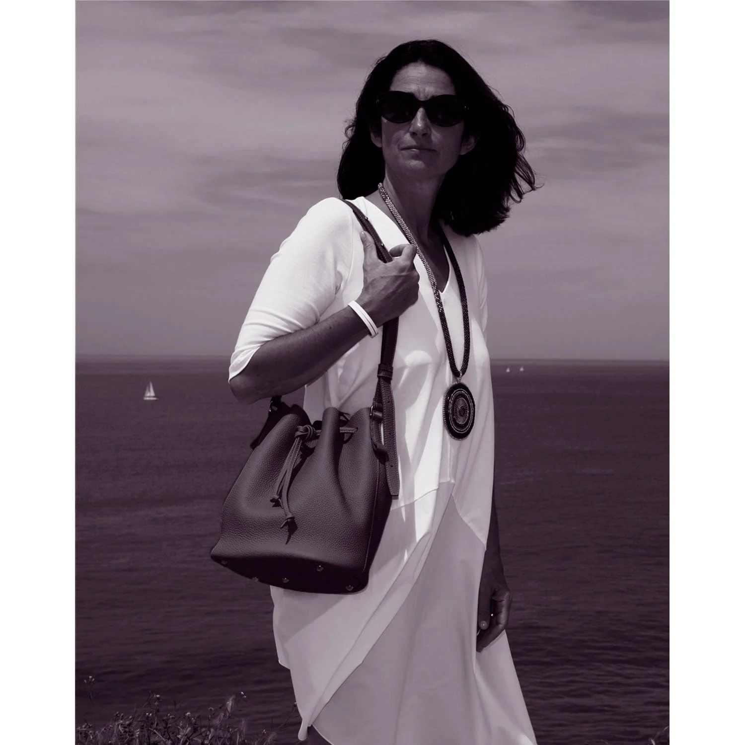 Une femme portant la robe LOUISON, accompagnée d'un sac à main et d'un collier, se tient au bord de l'océan avec des lunettes de soleil.