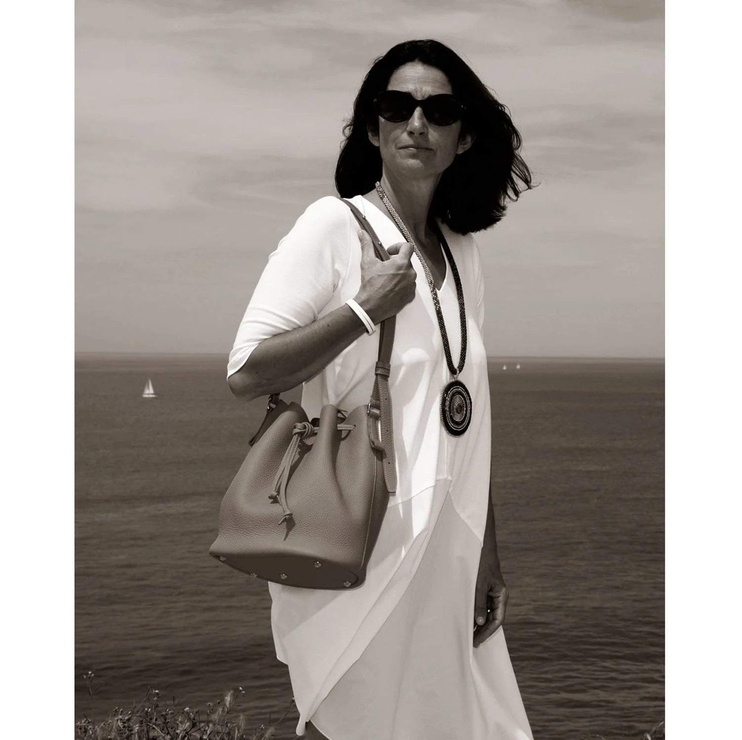 Une femme portant des lunettes de soleil et une robe blanche se tient au bord de l'océan, tenant le sac à main LOUISON et portant un long collier.