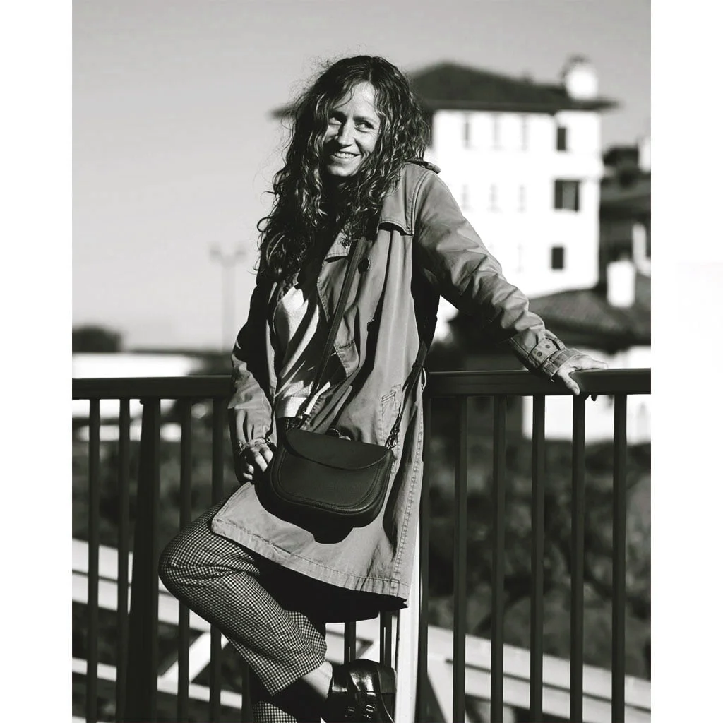 Une personne aux cheveux longs et portant un manteau se tient debout contre une balustrade, souriant comme si elle était portée disparue. Il y a un bâtiment en arrière-plan et l'image est en noir et blanc.