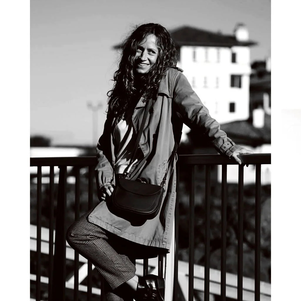 Photo en noir et blanc d'une personne souriante, appuyée contre une balustrade avec le sac à main MIA, devant un bâtiment flou en arrière-plan.