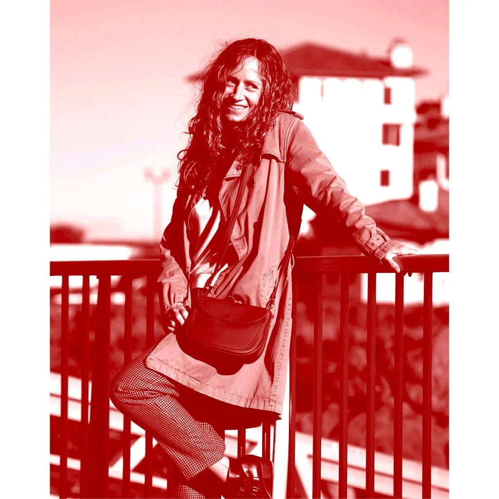 Une femme aux cheveux bouclés, vêtue du manteau MIA et portant un sac, s'appuie contre une balustrade en souriant. L'image présente une teinte rouge.