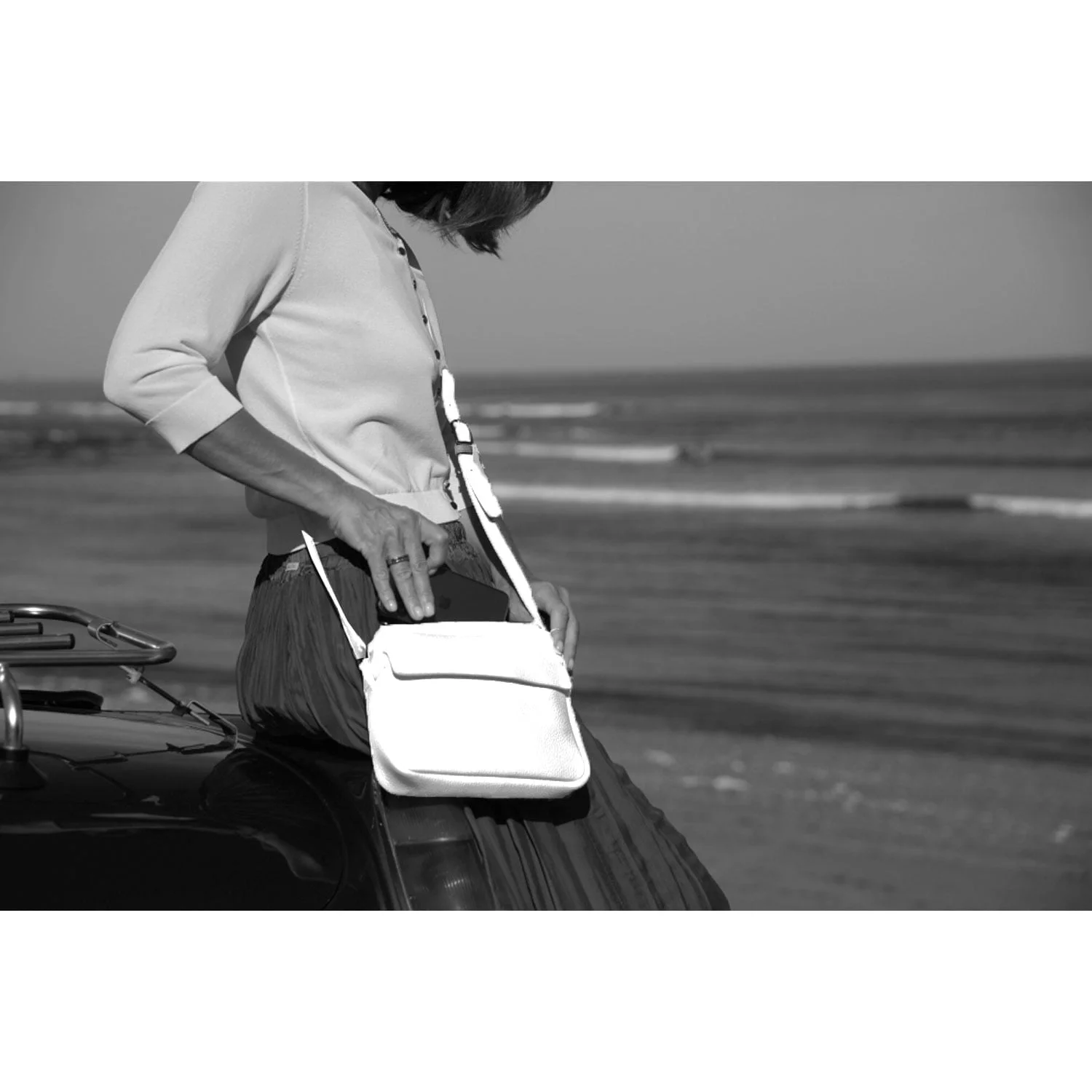 Une personne assise sur le toit d'une voiture, vêtue d'un pull et d'une jupe, tient le sac à bandoulière blanc ZOE avec l'océan en arrière-plan.