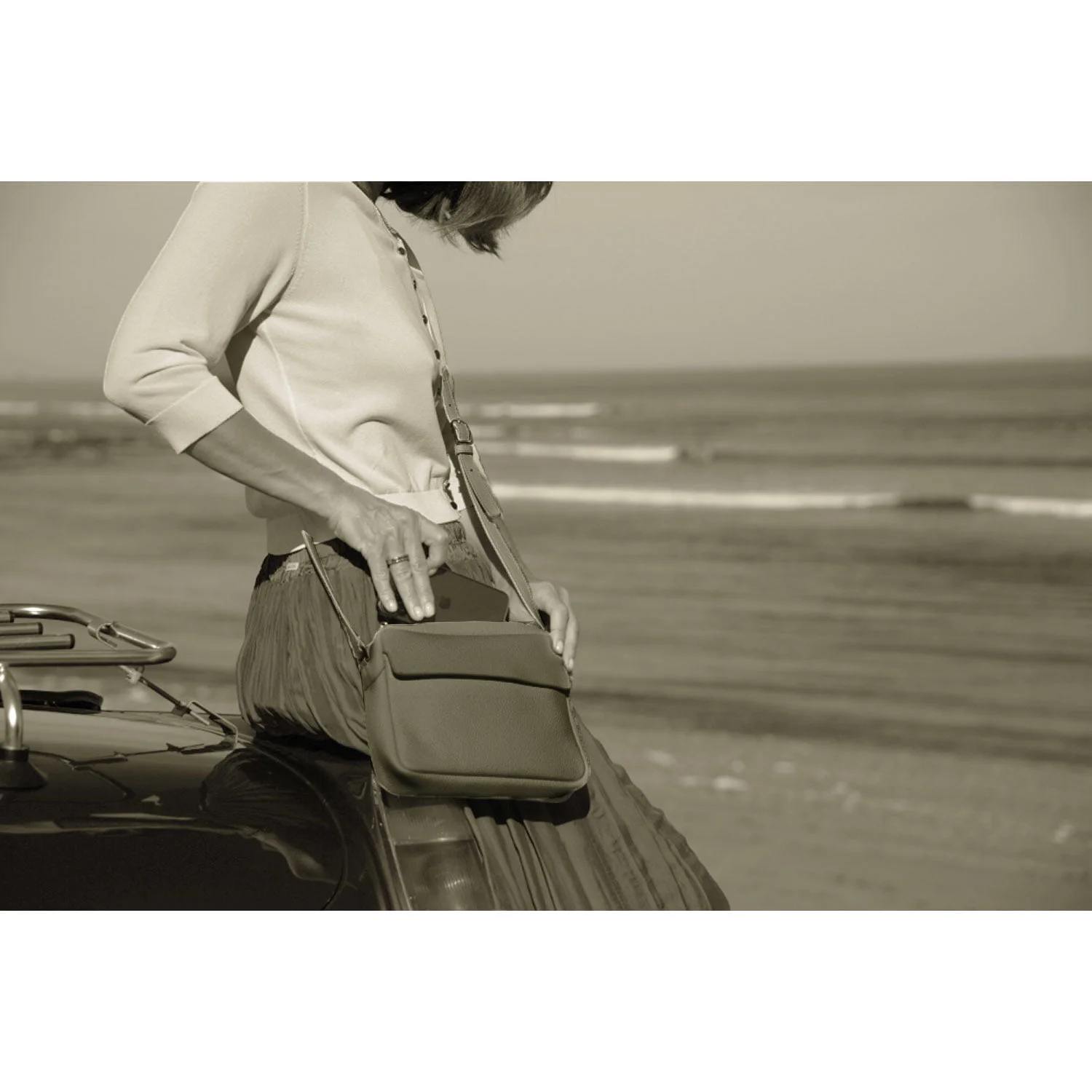 Personne assise sur une voiture au bord de la plage, portant une jupe longue et un pull, tenant le sac à bandoulière ZOE.