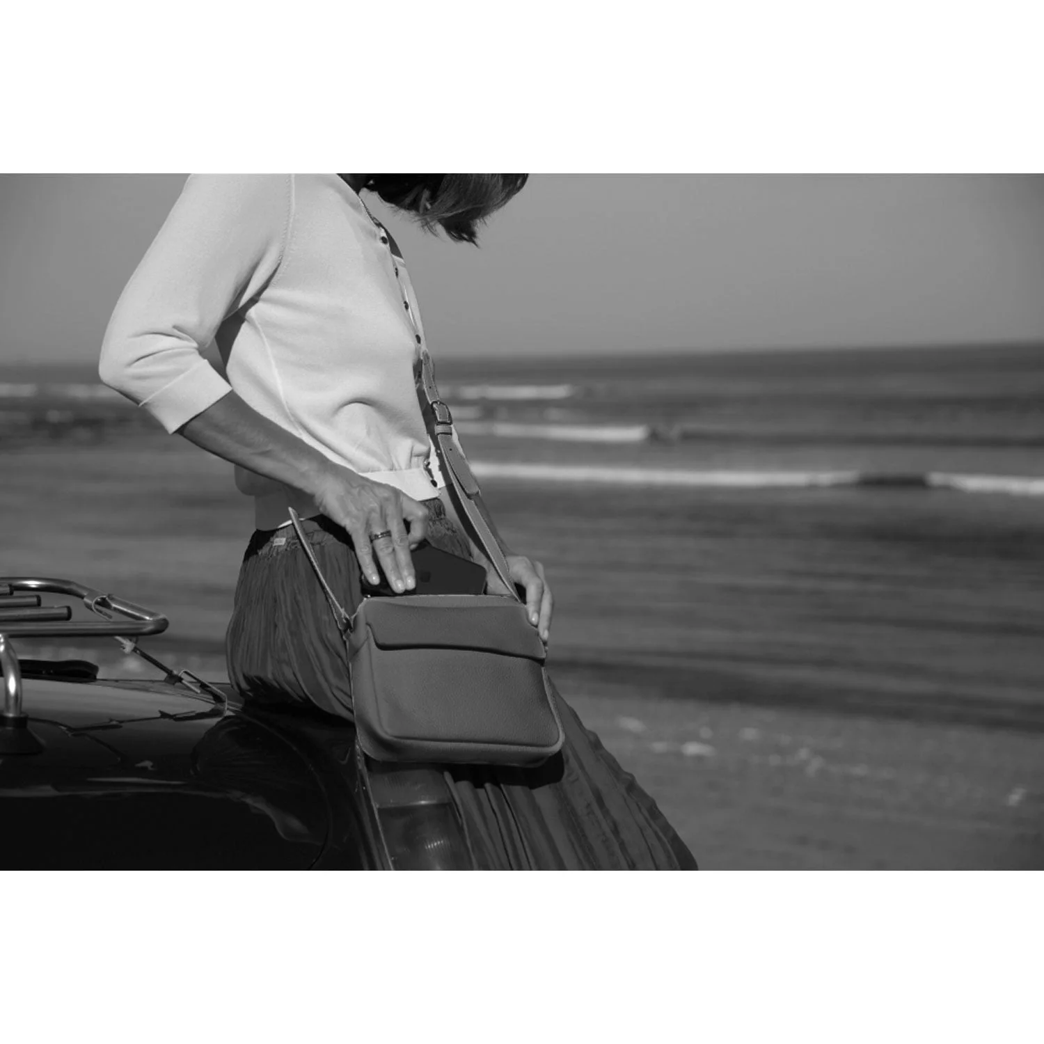 Une personne est assise sur une voiture au bord de la plage, vêtue d'un haut blanc et d'une jupe plissée, tenant le sac à bandoulière ZOE avec l'océan en arrière-plan. La scène est capturée en noir et blanc.