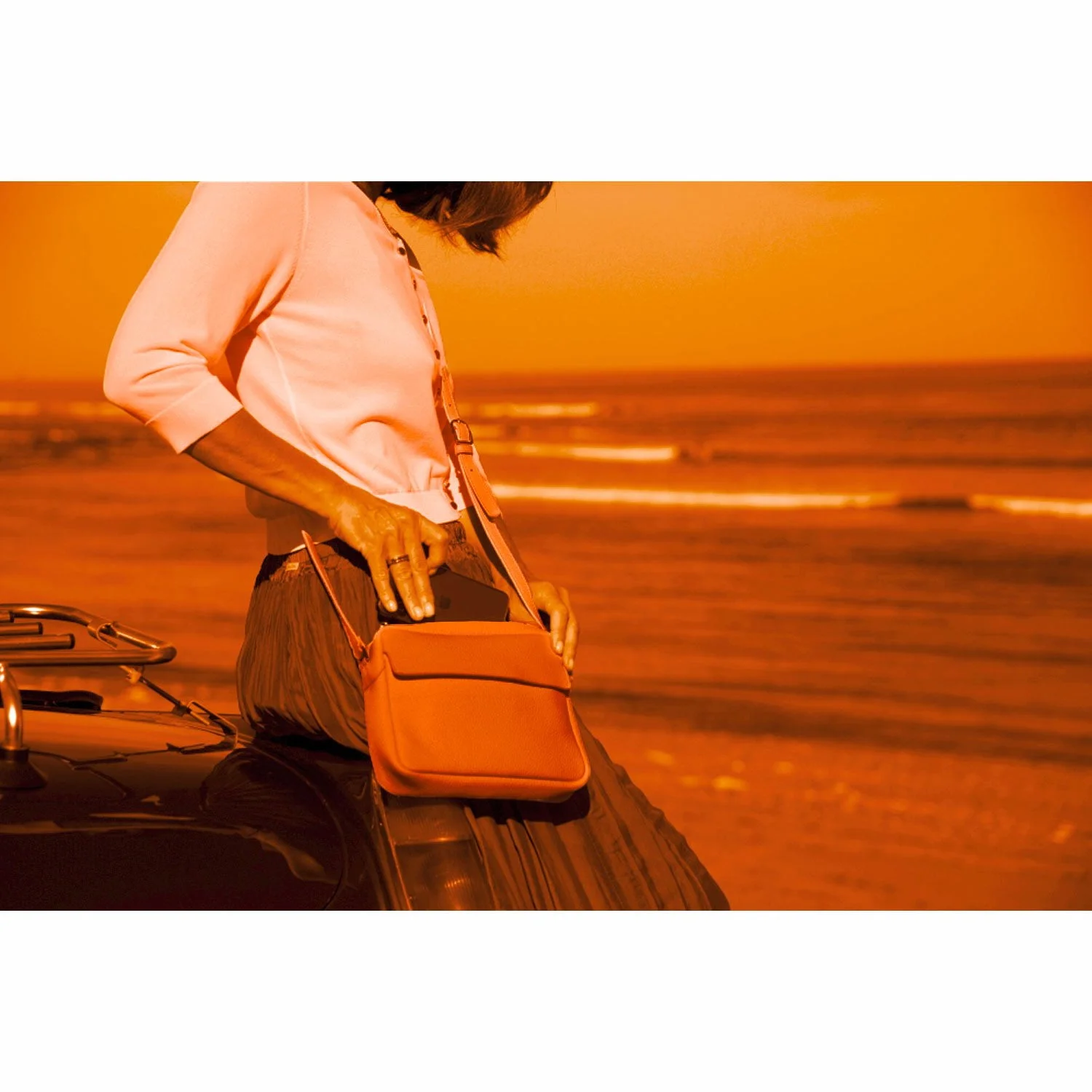 Une personne se tient debout à côté d'une voiture sur une plage avec des vagues en arrière-plan, plaçant un ordinateur portable ou un téléphone dans un sac à bandoulière orange ZOE. L'image est rehaussée d'un filtre orange chaud.