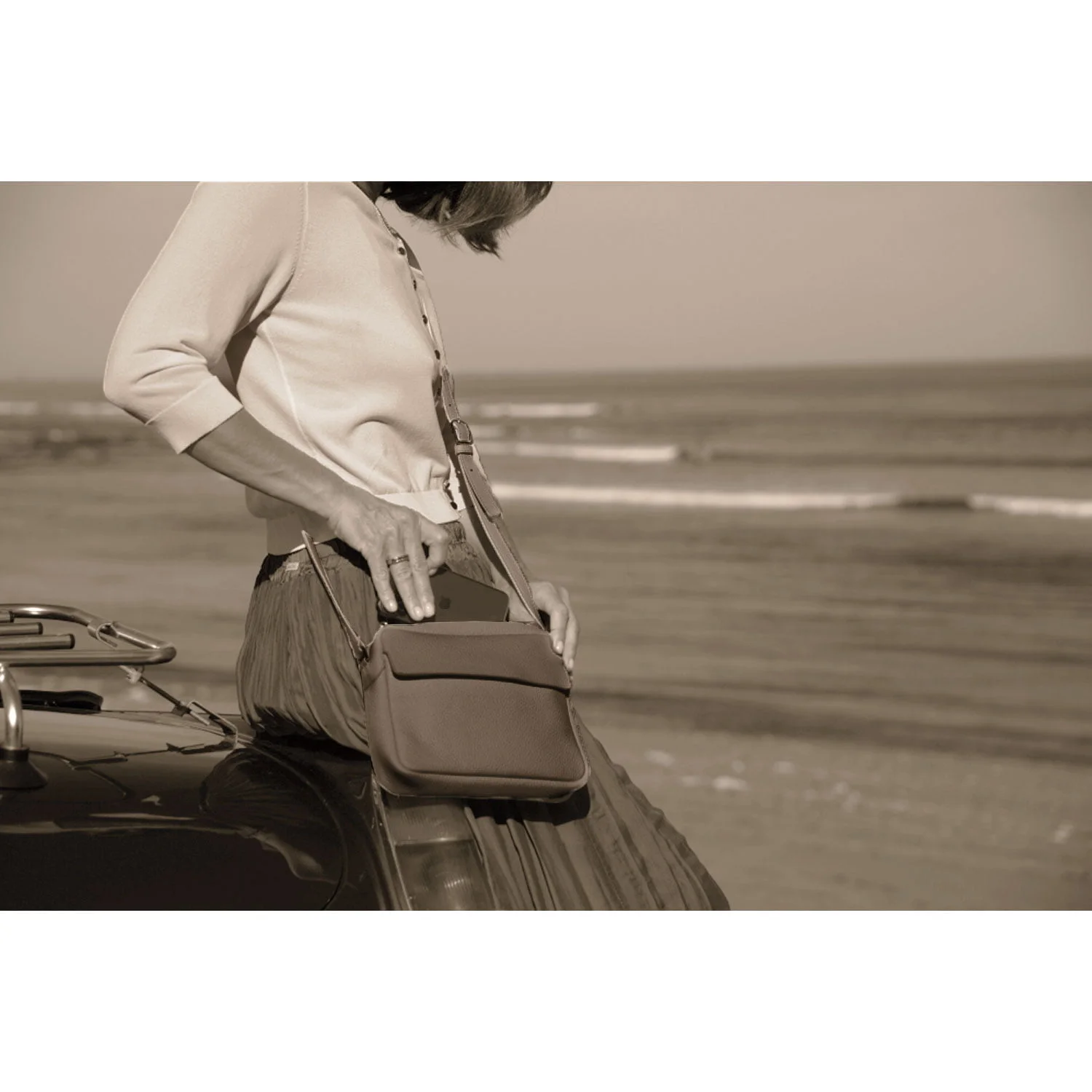 Une personne s'appuie contre une voiture sur la plage, en train d'ajuster son sac bandoulière ZOE. La photo est en sépia.