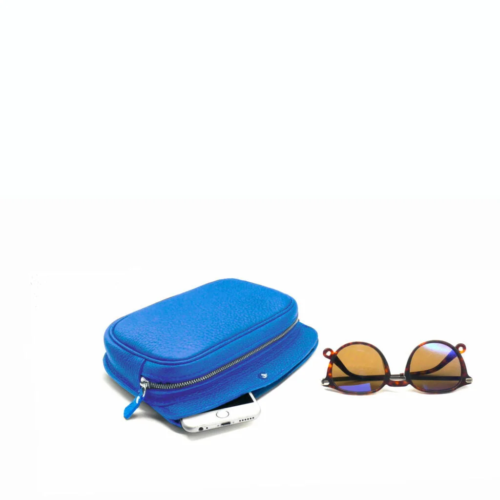 Pochette zippée bleue avec un smartphone partiellement à l'intérieur et des lunettes de soleil en écaille de tortue avec des verres teintés à côté, sur un fond blanc uni.
