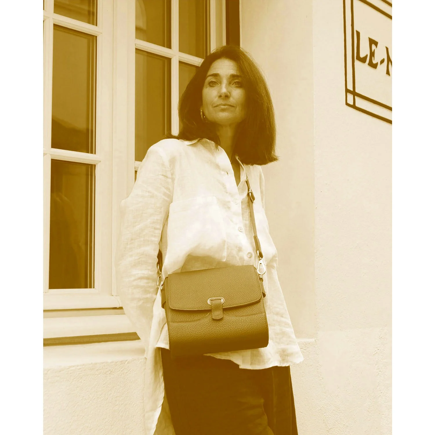 Une femme en chemise blanche s'appuie contre un bâtiment, portant le sac bandoulière SAM. Le mur présente un panneau partiellement visible et la photo est en sépia.