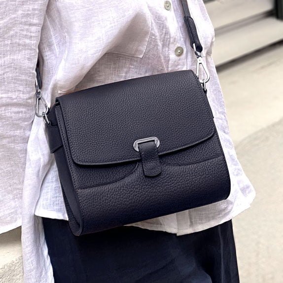 Une personne portant une chemise blanche et un pantalon noir porte un sac à main en cuir noir avec un fermoir argenté.