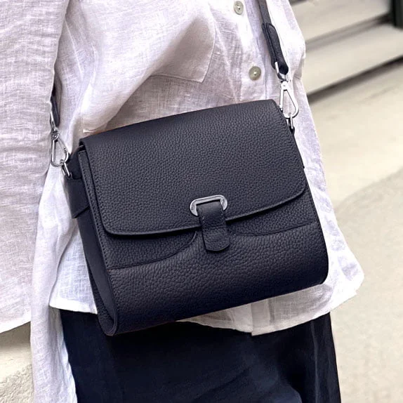 Une personne portant une chemise blanche et un pantalon noir porte un sac à main en cuir noir avec un fermoir argenté.