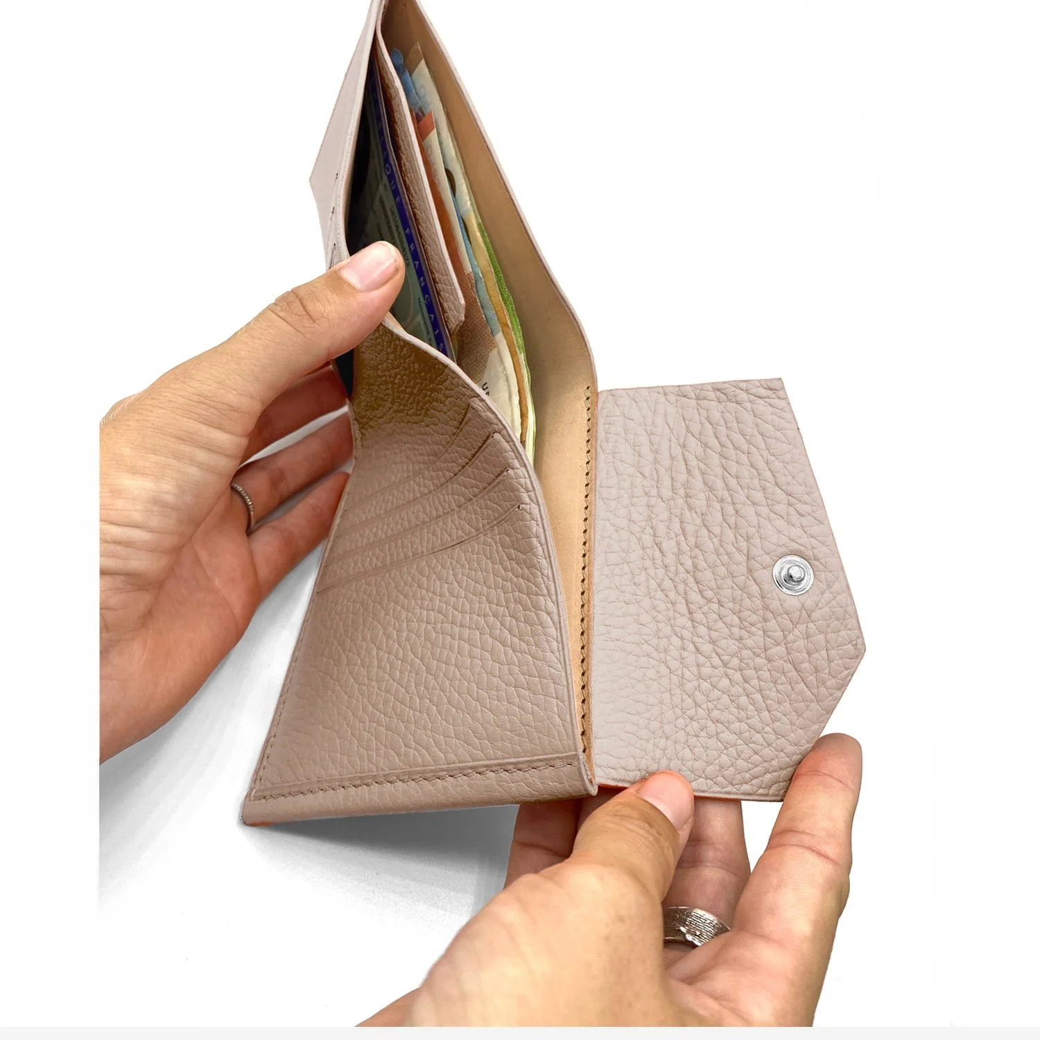Deux mains tiennent un portefeuille en cuir beige ouvert contenant de l'argent et des cartes, avec une fermeture à pression en métal visible.