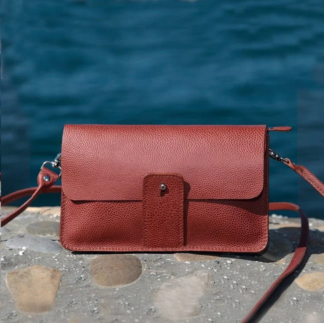 Sac bandoulière en cuir marron sur une surface en pierre avec de l'eau en arrière-plan.