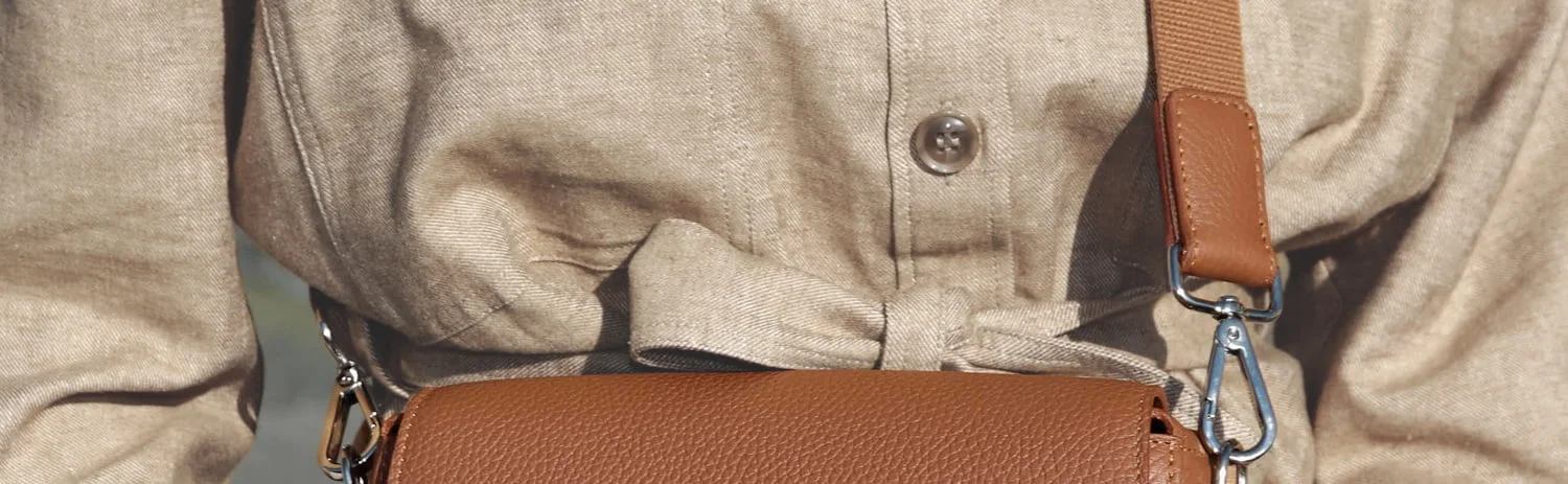 Gros plan d'une personne portant un manteau boutonné beige avec une taille nouée, accessoirisé d'un sac bandoulière en cuir marron.