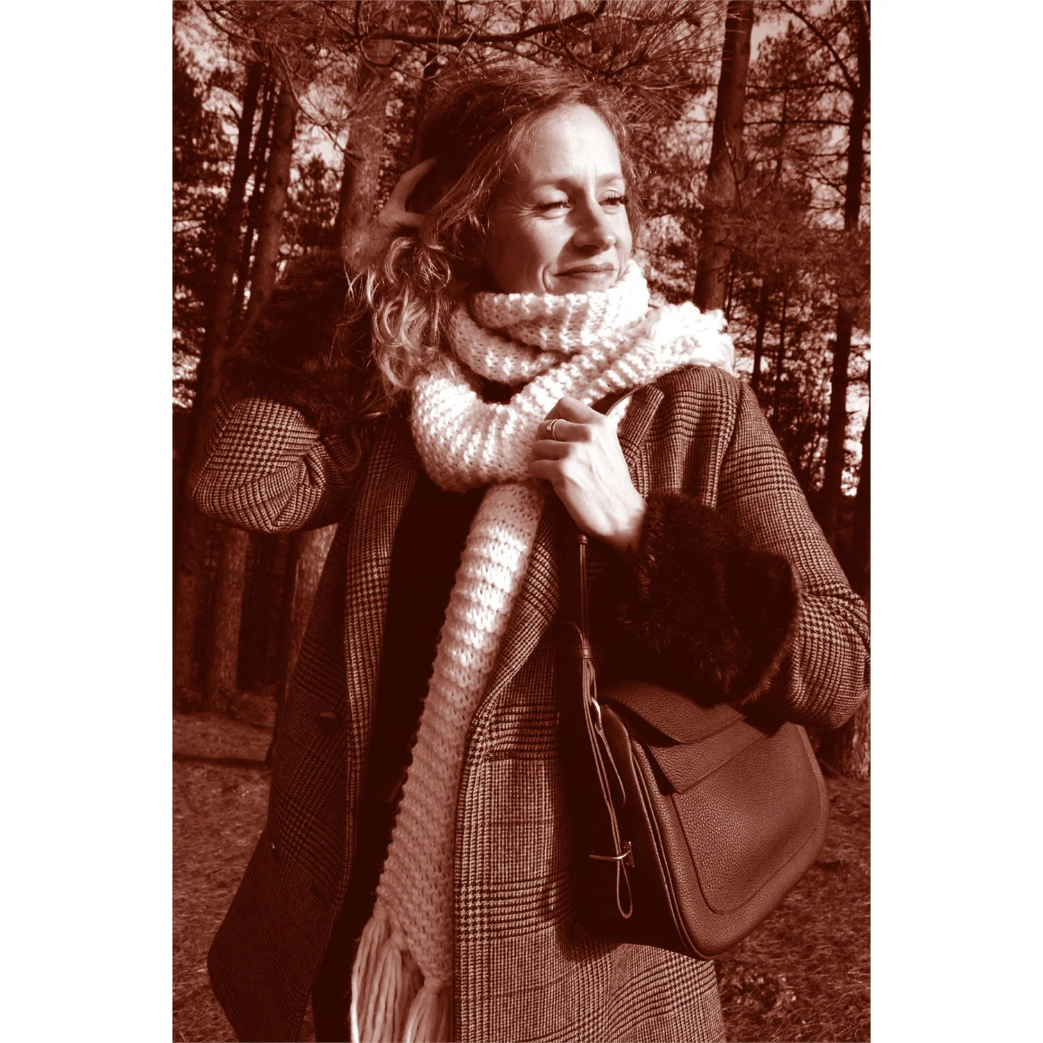 Une femme aux cheveux bouclés, portant un manteau et un foulard, se tient debout dans une zone boisée, tenant un sac et regardant sur le côté. L'image est en ton sépia.