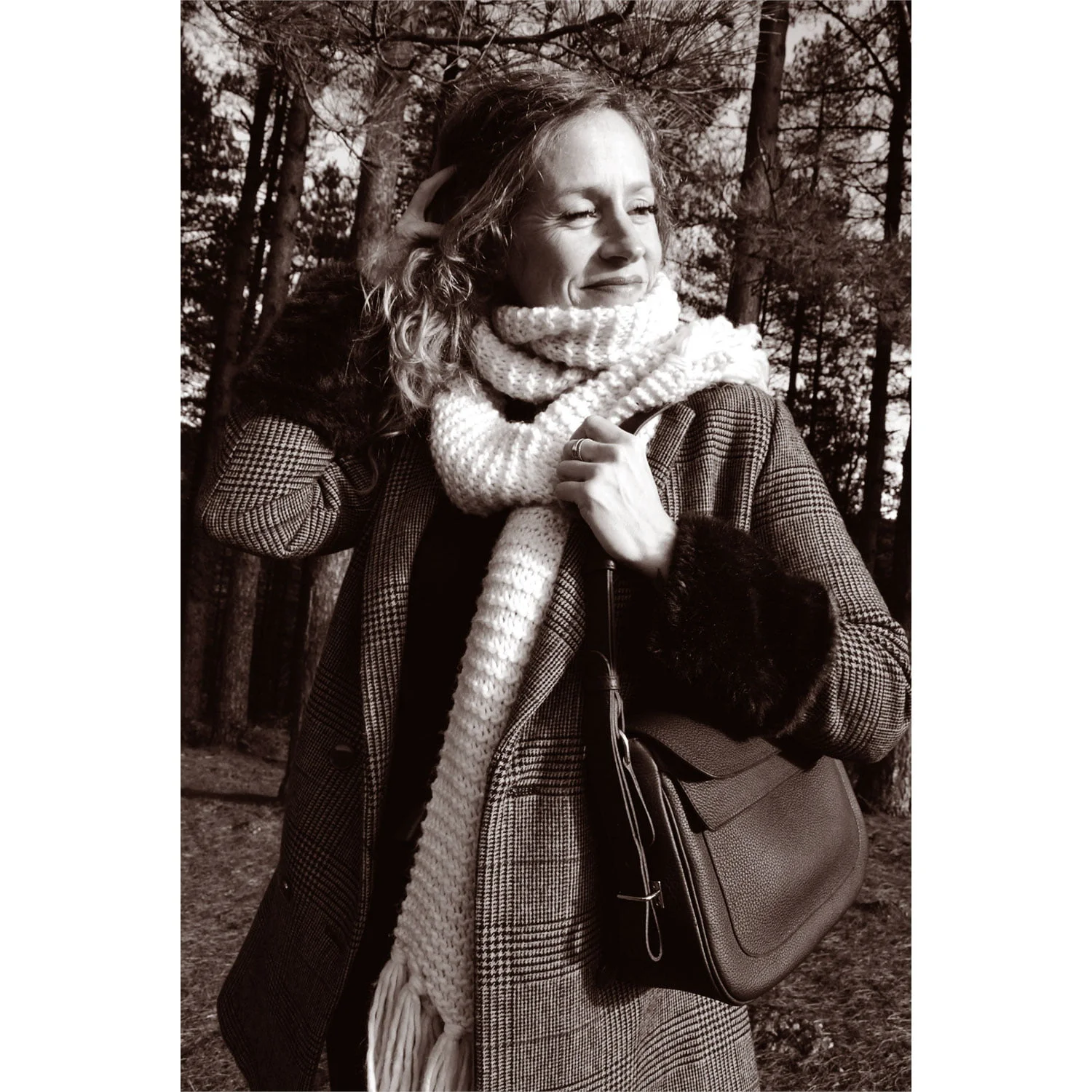 Une femme portant un manteau à carreaux et une écharpe se tient dehors dans une zone boisée, tenant un sac et souriant tout en regardant sur le côté. Photo en noir et blanc.