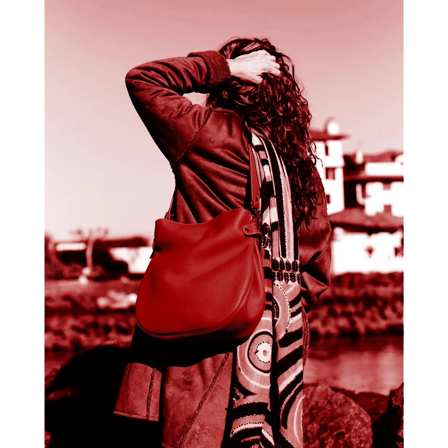 Une personne aux cheveux longs et bouclés, portant un sac à main et un manteau et un foulard à motifs, se tient debout près de l'eau, le dos tourné. L'image est teintée de rouge.
