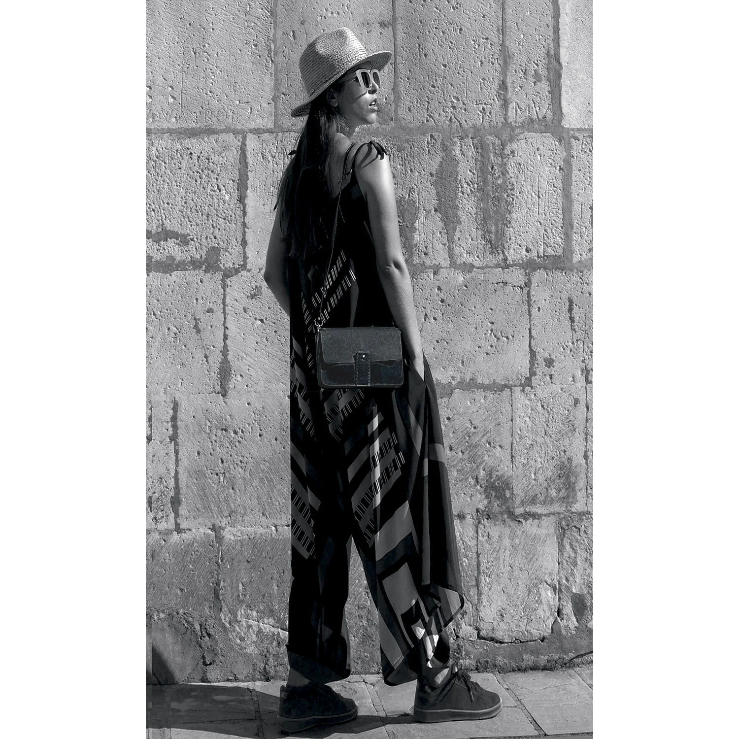 Une femme portant un chapeau et des lunettes de soleil se tient devant un mur de pierre, vêtue d'une longue robe à motifs et tenant une pochette.