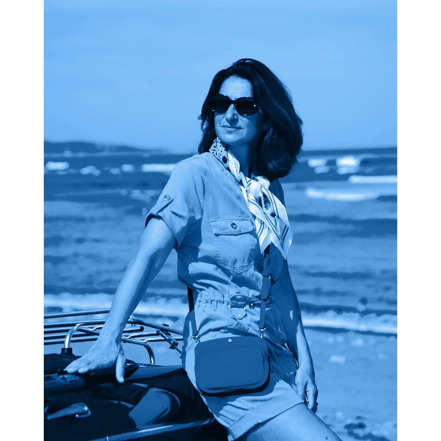 Une femme portant des lunettes de soleil s'appuie sur une voiture près de la plage. Elle porte un foulard, une tenue à manches courtes et un petit sac. L'image est teintée de bleu.
