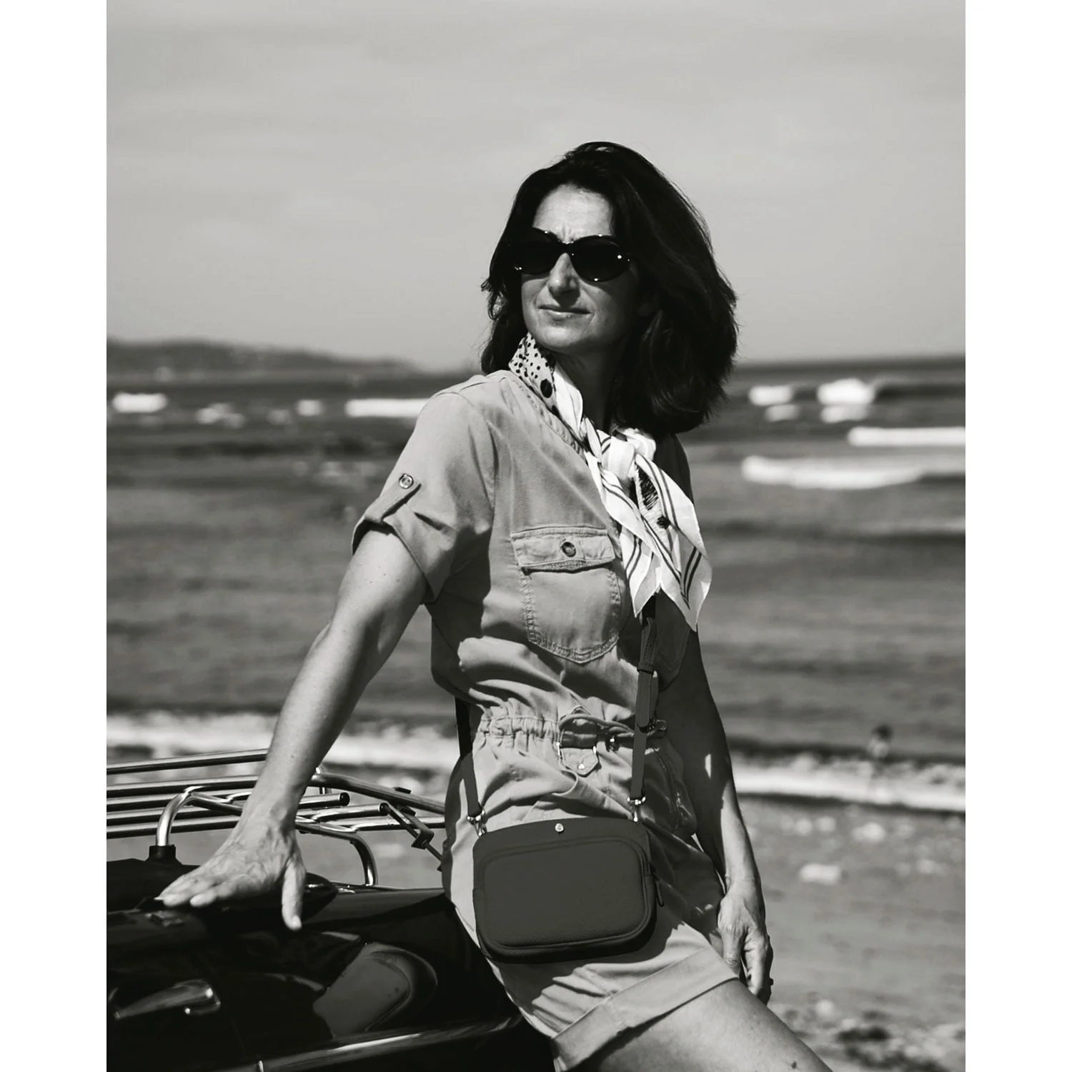 Une femme portant des lunettes de soleil s'appuie sur une voiture au bord de la plage, portant un foulard et une robe ceinturée. Les vagues de l'océan sont visibles en arrière-plan.