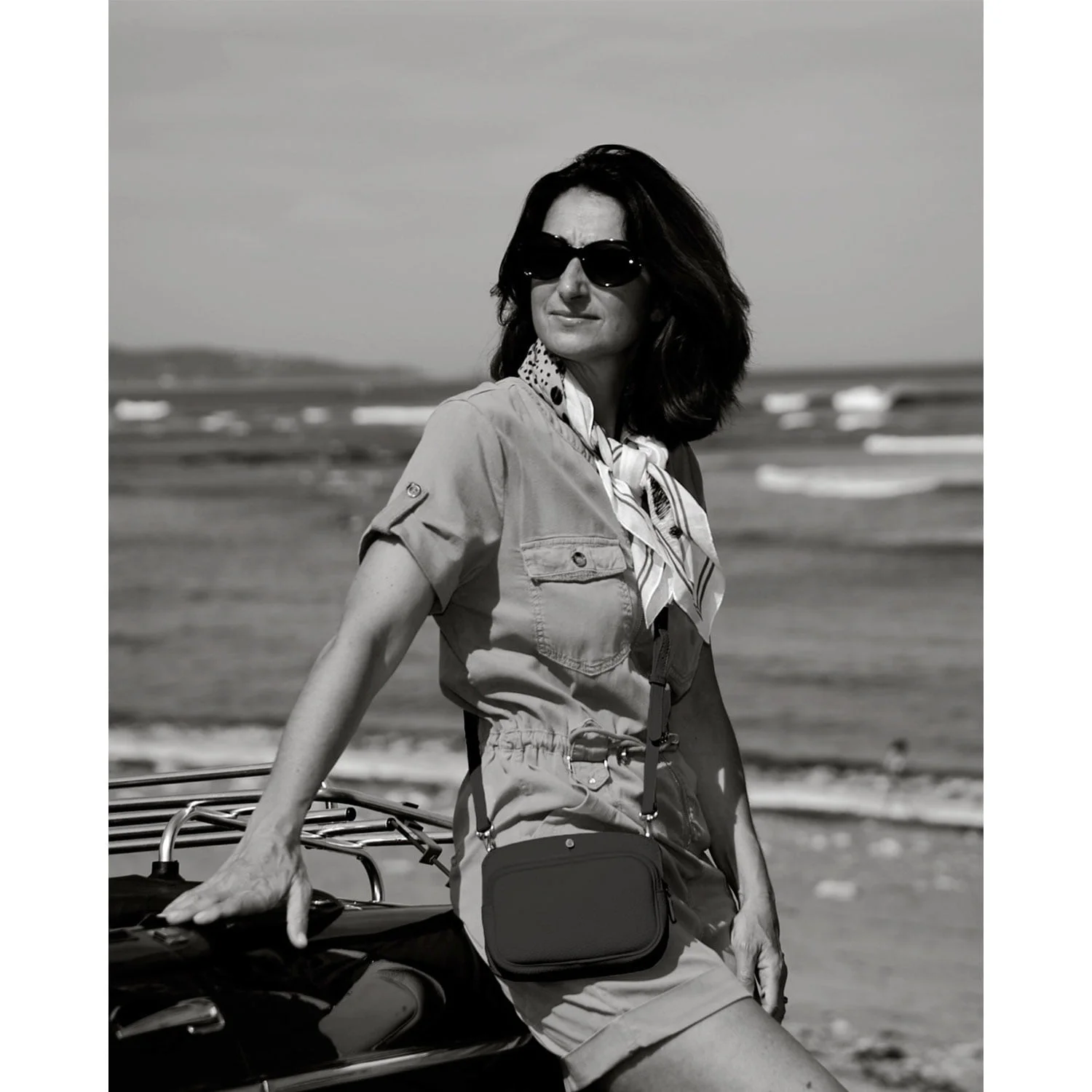 Une femme portant des lunettes de soleil et un foulard s'appuie sur une voiture près de la plage, le regard perdu au loin. La scène est en noir et blanc.
