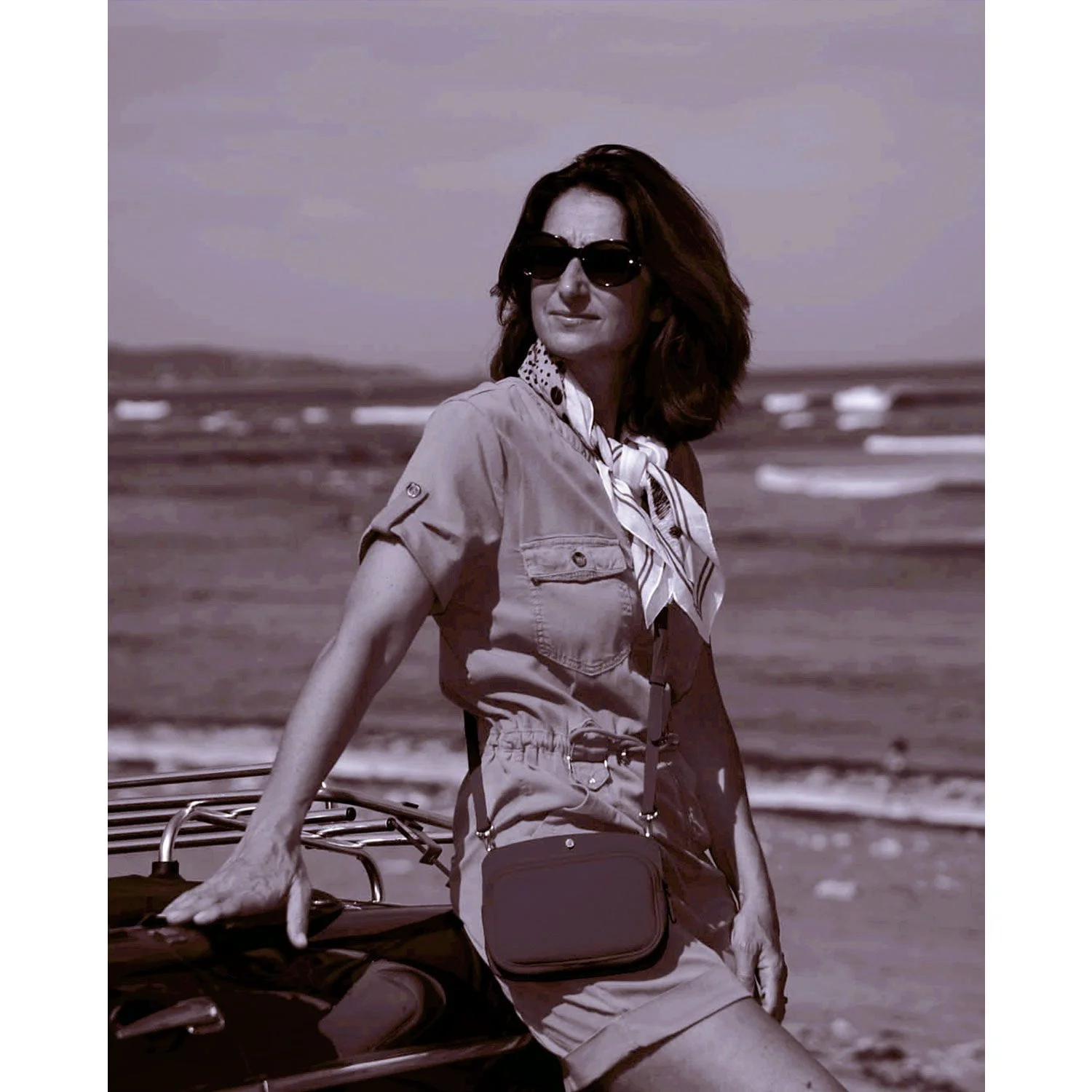 Une femme portant des lunettes de soleil et un foulard se tient près d'une voiture sur une plage avec des vagues en arrière-plan.