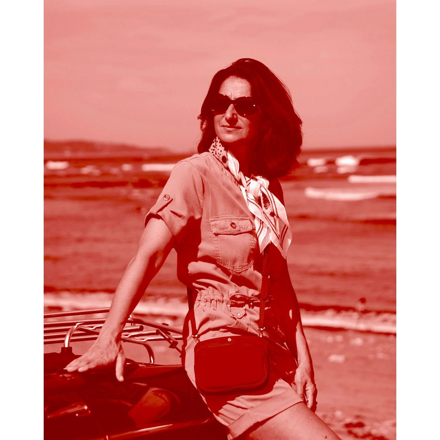 Une femme portant des lunettes de soleil et un foulard à motifs se tient près d'une voiture sur la plage, face à la mer. L'image a une teinte rouge.