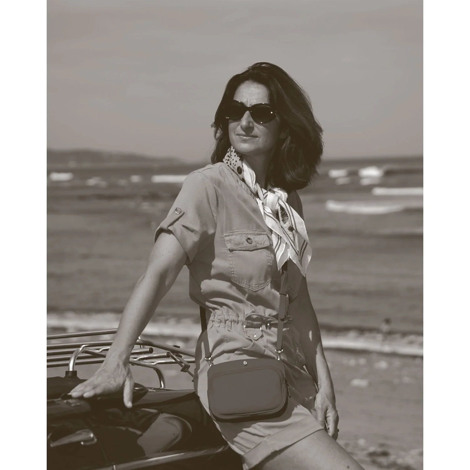 Une femme portant des lunettes de soleil s'appuie contre une voiture près de la plage, vêtue d'une tenue décontractée avec foulard et sac. Photo de style vintage.