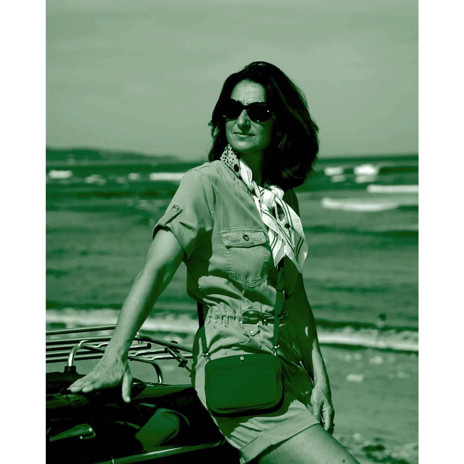 Une femme portant des lunettes de soleil se tient près d'une voiture près de la plage, vêtue d'une robe et d'un foulard.
