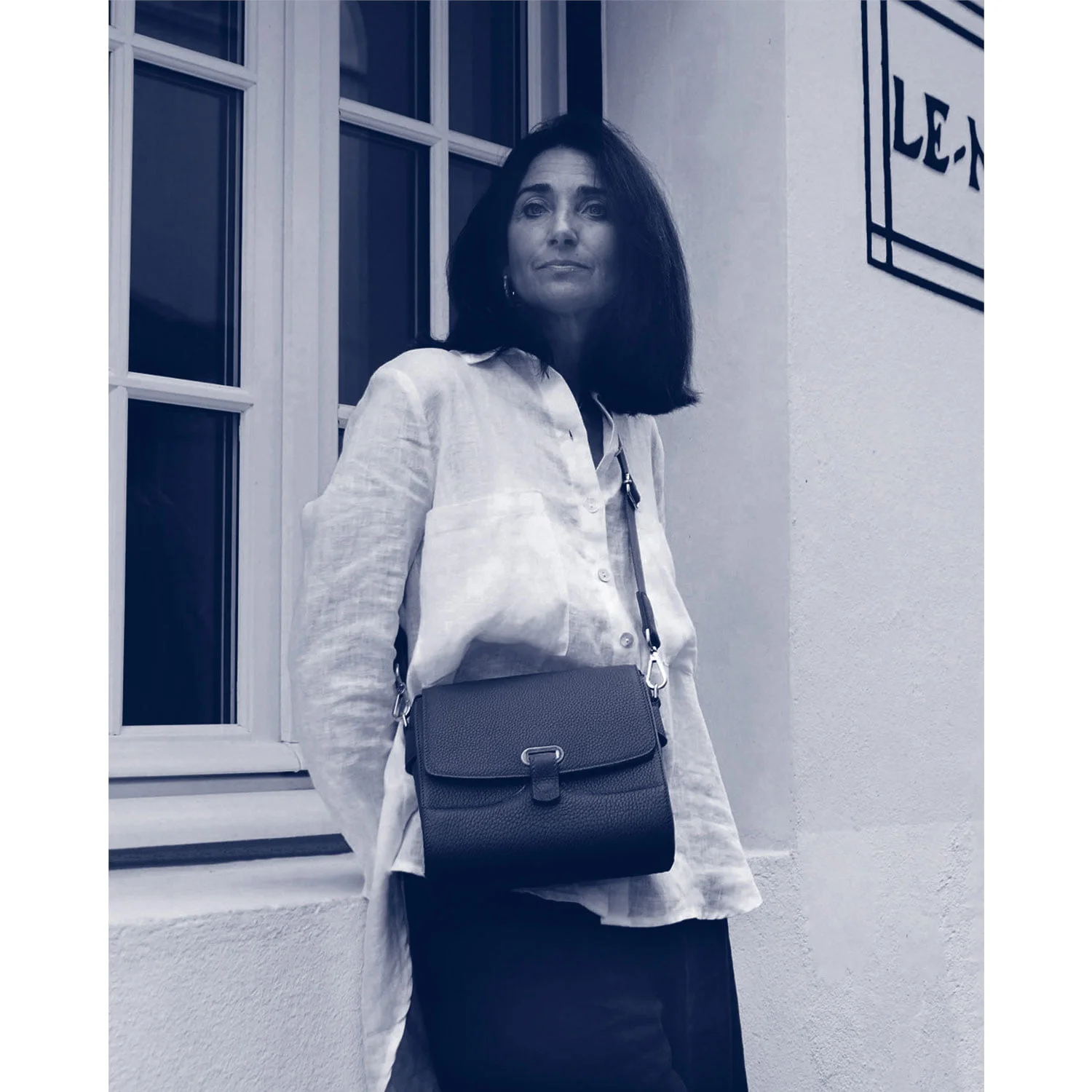 Une personne aux cheveux mi-longs se tient près d'une fenêtre, portant une chemise de couleur claire et un sac à bandoulière foncé.