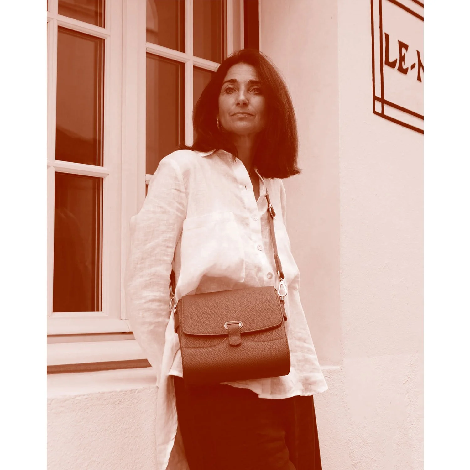 Une personne aux cheveux mi-longs, portant un chemisier de couleur claire et un sac, se tient près d'une fenêtre et d'un mur.