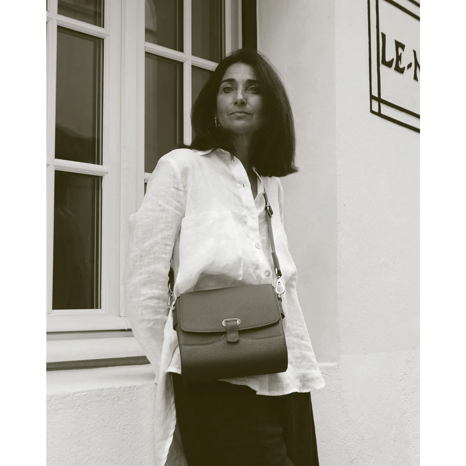 Une femme portant un chemisier blanc et un sac à bandoulière se tient contre un mur près d'une fenêtre.