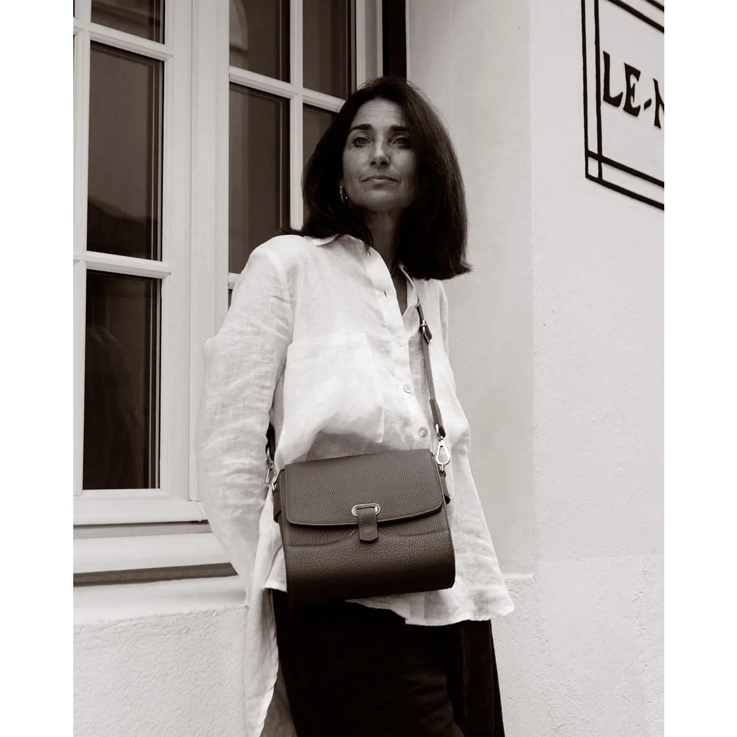 Une femme en chemise blanche se tient près d'une fenêtre avec un sac à bandoulière, regardant devant elle.