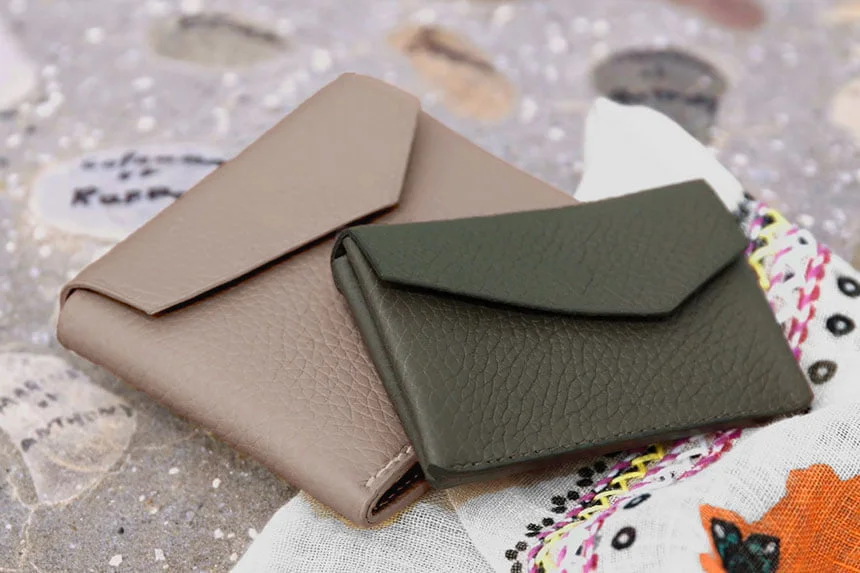 Deux portefeuilles en cuir texturé, l'un beige et l'autre vert, reposent sur un tissu à motifs posé sur une surface sablonneuse avec des coquillages.