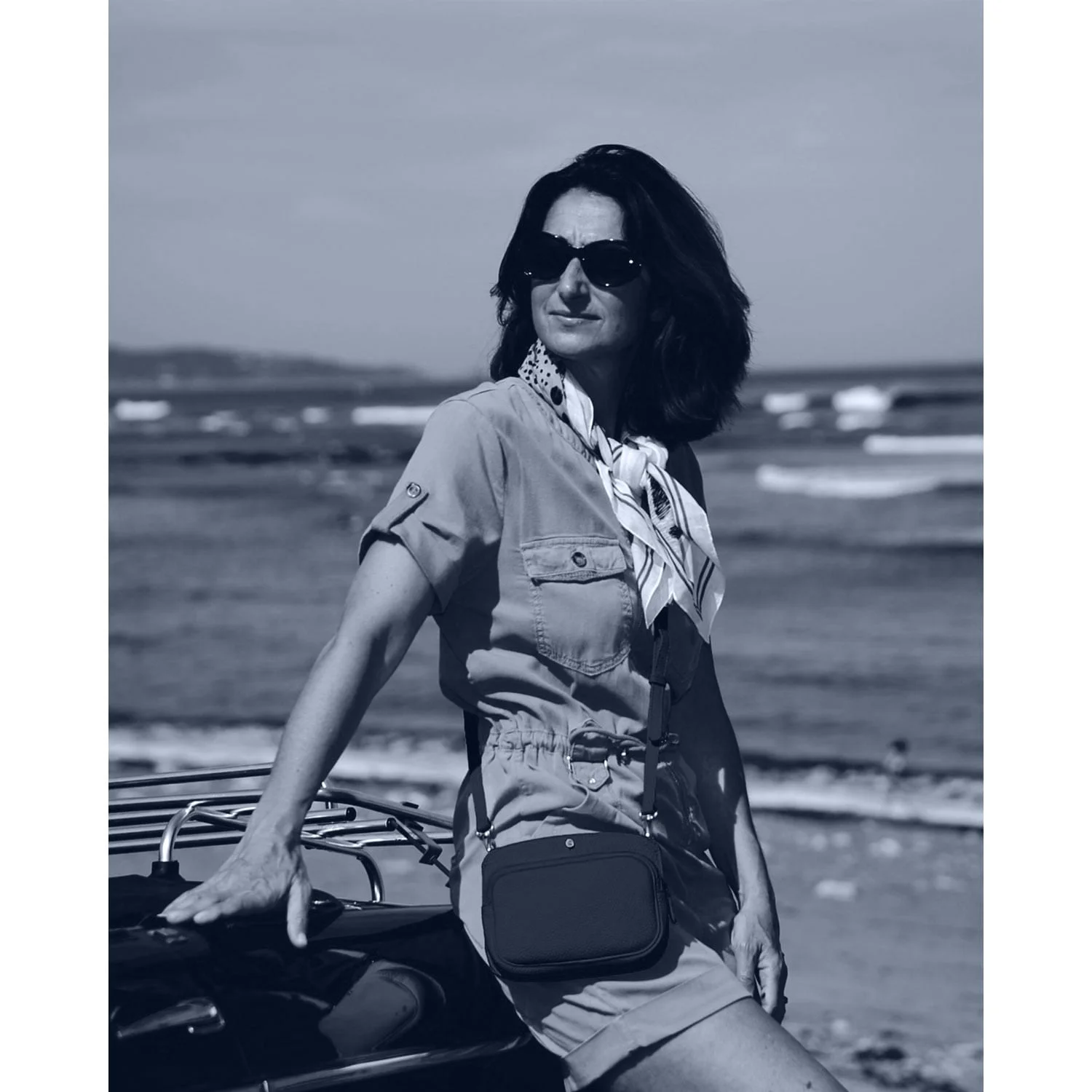Une femme portant des lunettes de soleil et un foulard s'appuie contre une voiture sur une plage. Elle porte une robe ceinturée et porte un petit sac à la taille. On peut voir des vagues en arrière-plan.