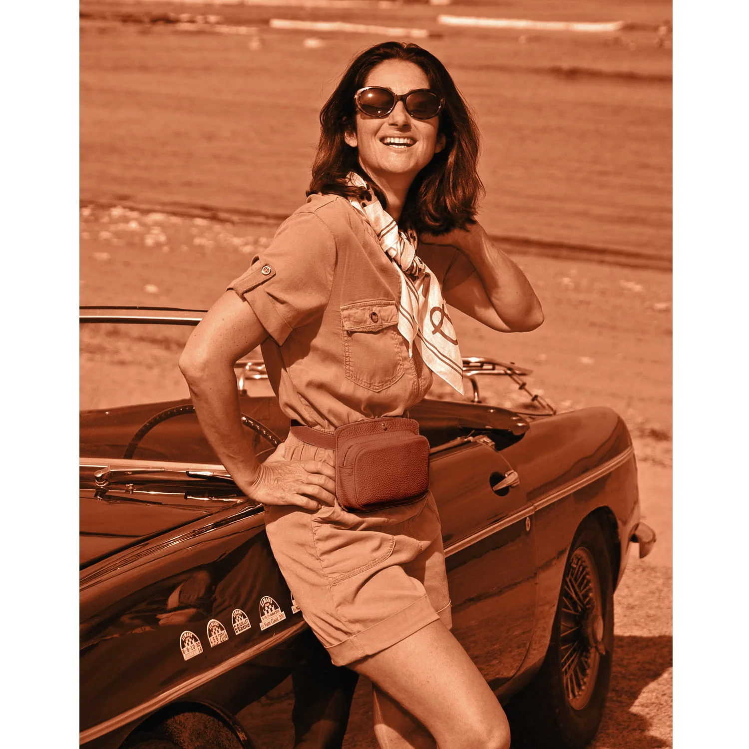 Une femme portant des lunettes de soleil et un foulard s'appuie sur une voiture vintage près de la plage, souriant.