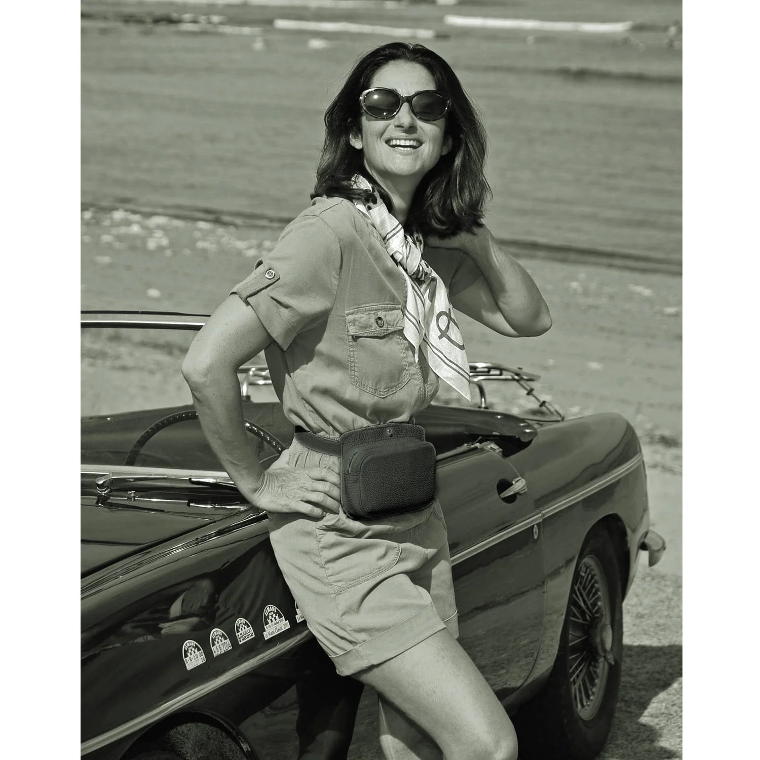 Une femme portant des lunettes de soleil se tient à côté d'une voiture ancienne sur la plage, portant un foulard et un sac banane.
