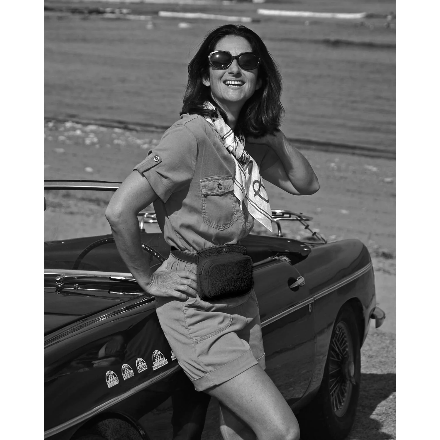 Une femme portant des lunettes de soleil et un foulard sourit tout en s'appuyant sur une voiture ancienne au bord de la plage. L'image est en noir et blanc.