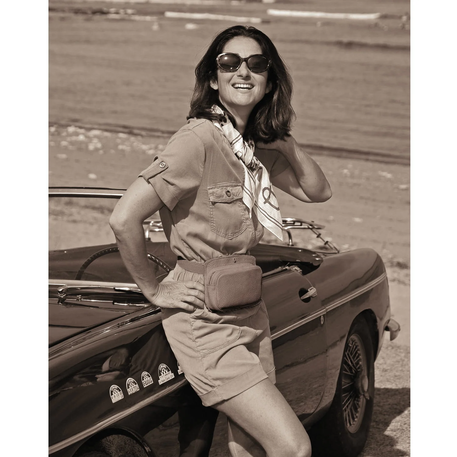 Une femme portant des lunettes de soleil et un foulard se tient à côté d'une voiture ancienne sur une plage.